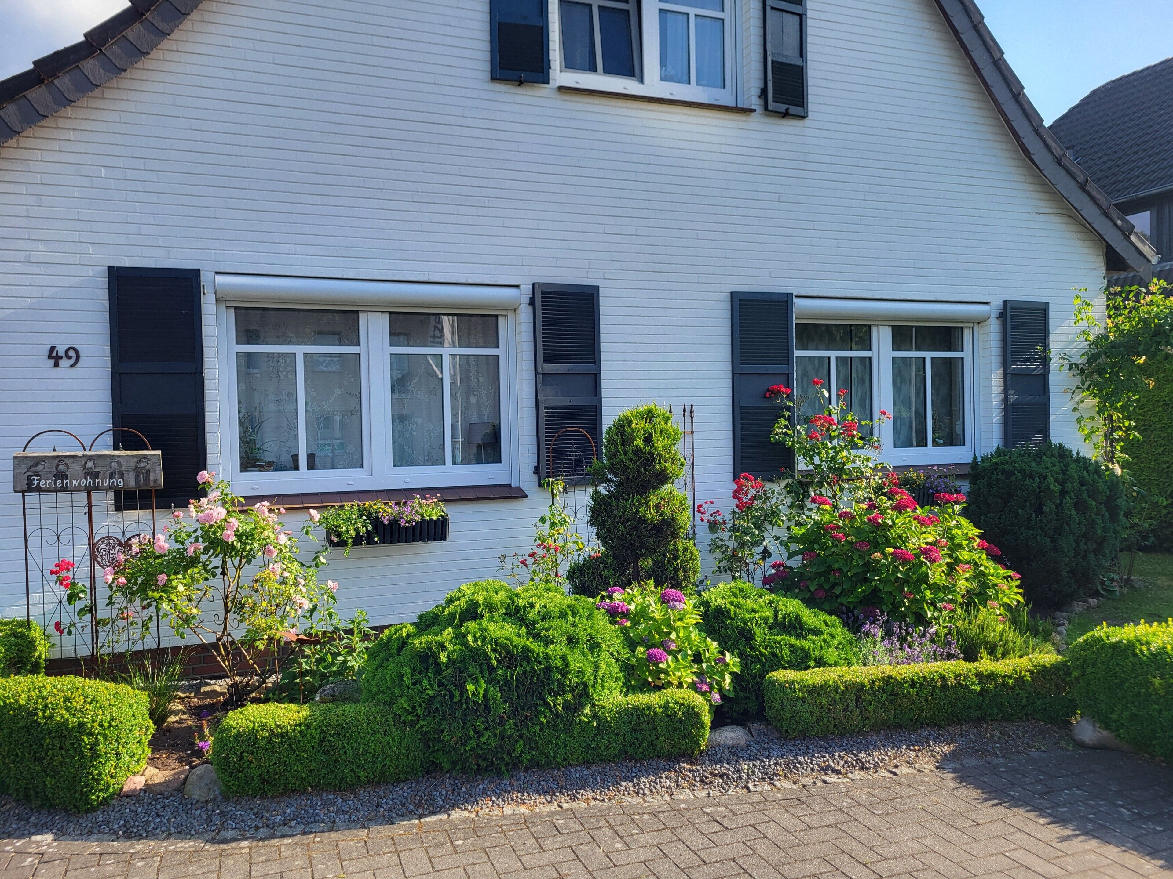 Un bâtiment blanc avec des volets noirs, une enseigne indiquant « Ferienwohnung 49 », et des jardins bien entretenus où poussent diverses plantes et fleurs.