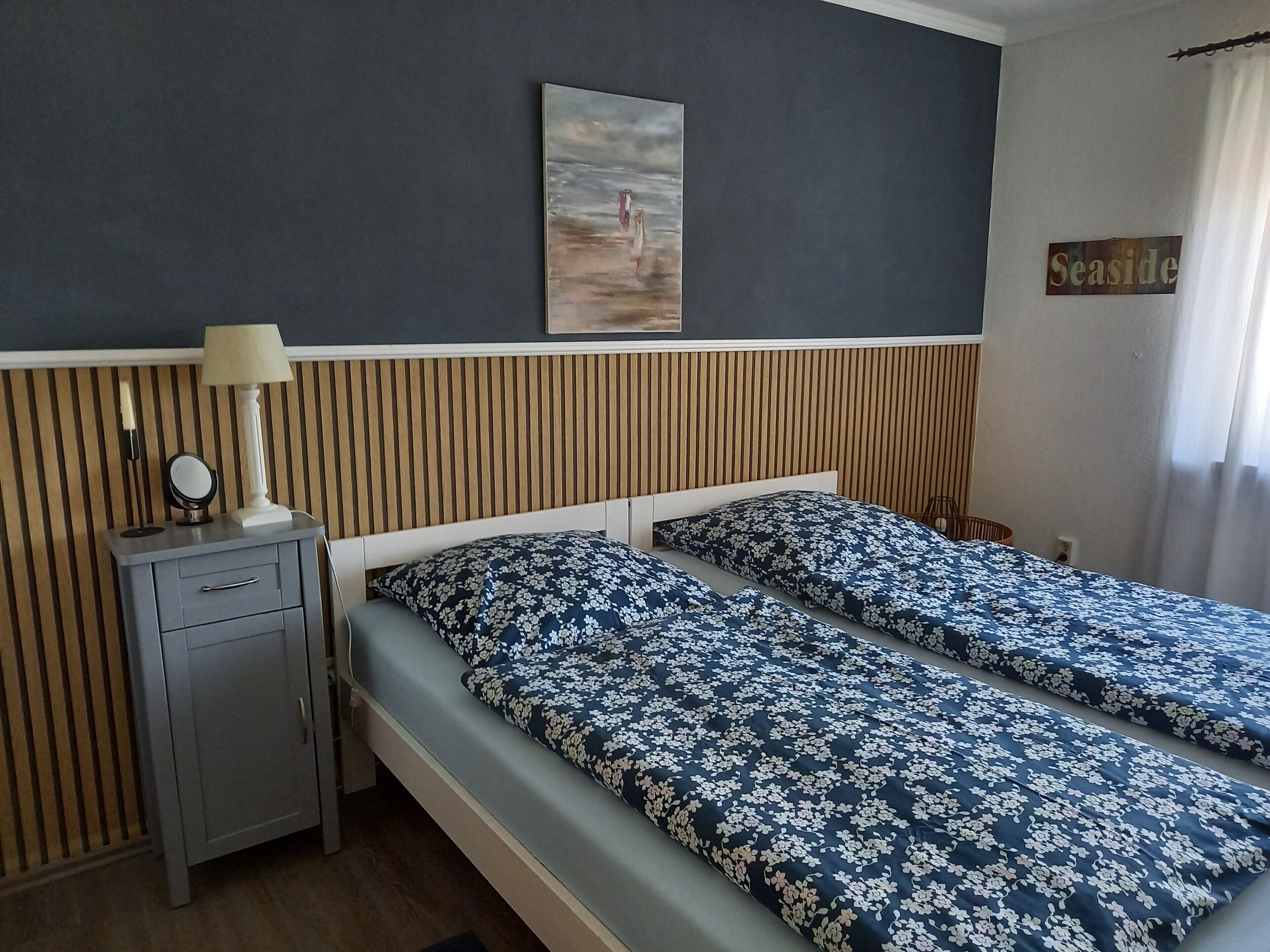 Uma cama de casal com roupa de cama floral, uma mesinha de cabeceira com abajur, um espelho pequeno e um quadro na parede.