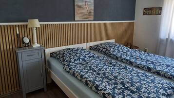 Uma cama de casal com roupa de cama floral, uma mesinha de cabeceira com abajur, um espelho pequeno e um quadro na parede.