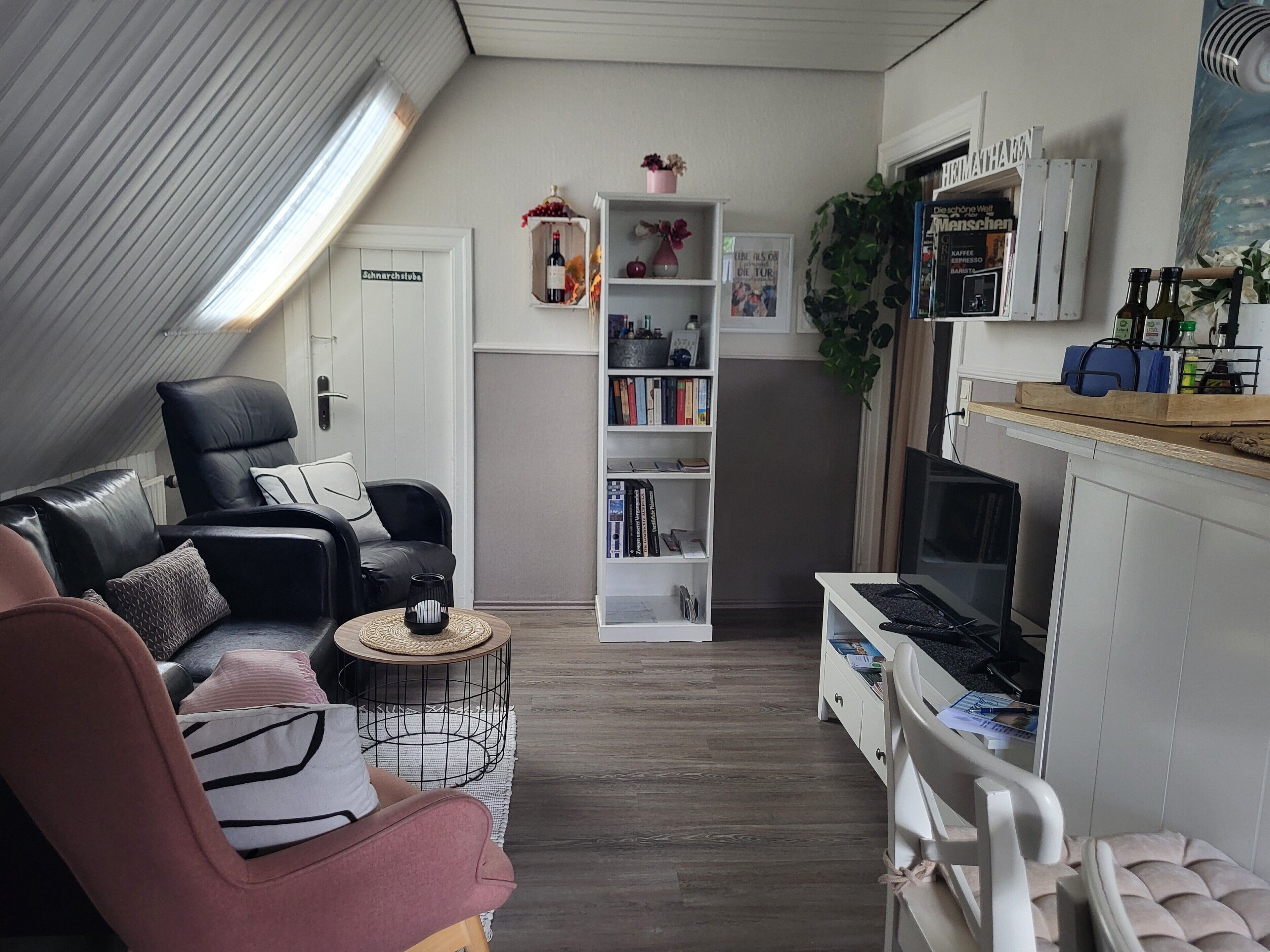 Una sala de estar acogedora con un sofá de cuero negro, un sillón rosa, una estantería blanca y un televisor sobre un soporte de madera.