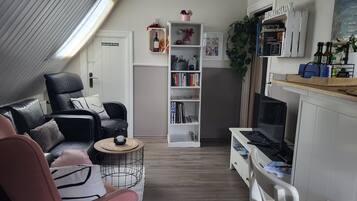 Una sala de estar acogedora con un sofá de cuero negro, un sillón rosa, una estantería blanca y un televisor sobre un soporte de madera.