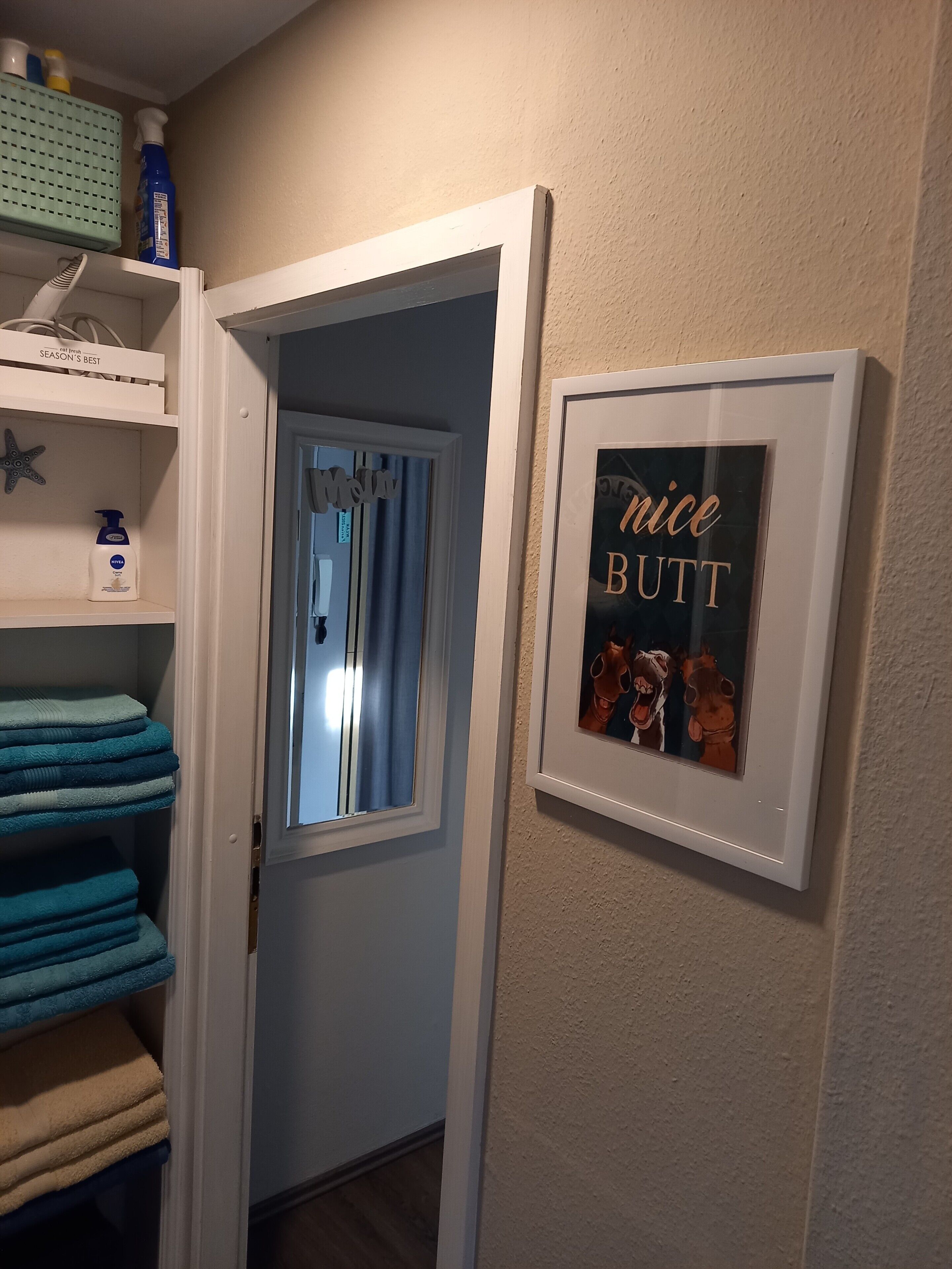 Ein Flur mit einer zurueckfuehrenden Fuehrung zu einem Zimmer, einem gerahmten Bild an der Wand und einer Ablage mit Handtuechern und Pflegeprodukten.
