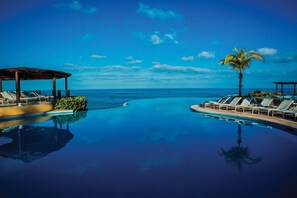 Una piscina con lettini e una palma, circondata da cielo azzurro e vista sull'oceano.