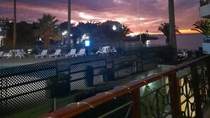 Ein Balkon mit Blick auf einen Sonnenuntergang über dem Meer, Palmen und einem Geländer mit Stühlen.