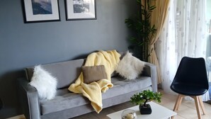 Ein Wohnzimmer mit grauem Sofa, weißem Couchtisch und schwarzem Sessel. An der Wand hängen zwei gerahmte Bilder, eine Topfpflanze und ein Fenster mit Vorhängen.