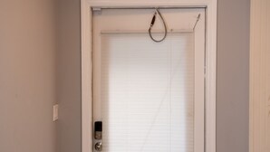 Une porte fermée surmontée d’une caméra de surveillance, une petite fenêtre avec des stores et un seuil blanc.