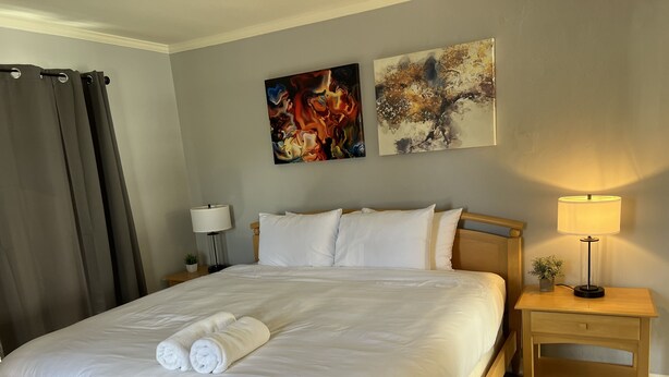 Un lit bien fait, avec du linge de lit blanc et des serviettes pliées, une tête de lit en bois, une lampe de chevet et deux tableaux abstraits accrochés au mur.