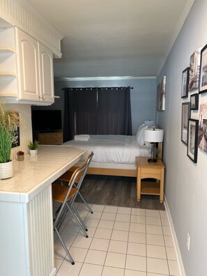A compact hotel room with a bed, a small desk, a chair, a nightstand, a TV, and a wall with framed pictures. - Studio- Sleep 2,Pool, Parking,5min walk to Beach 3 (Fort Lauderdale)