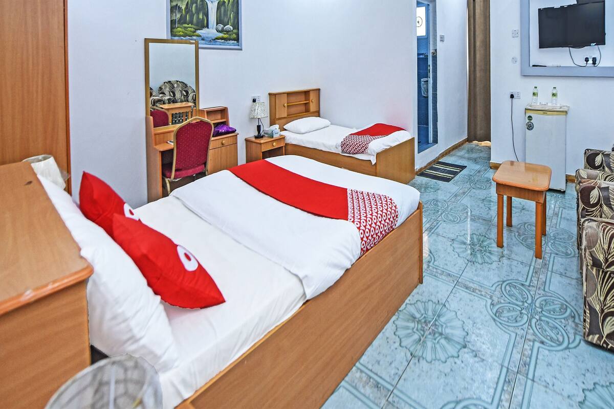 A hotel room with two beds, a desk, a chair, a small table, a mirror, a TV, and a radiator.
