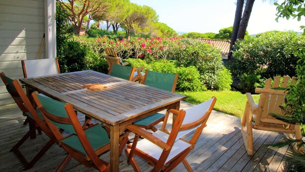 Una terraza de madera con una mesa y sillas, rodeada de abundante vegetación y vista al mar.