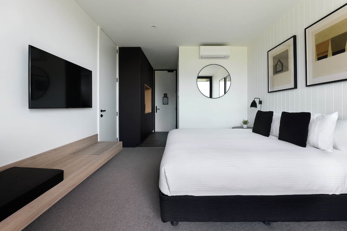 Una habitación de hotel moderna con una cama grande, un televisor de pantalla plana, una mesita de noche, un espejo y obras de arte enmarcadas en la pared.