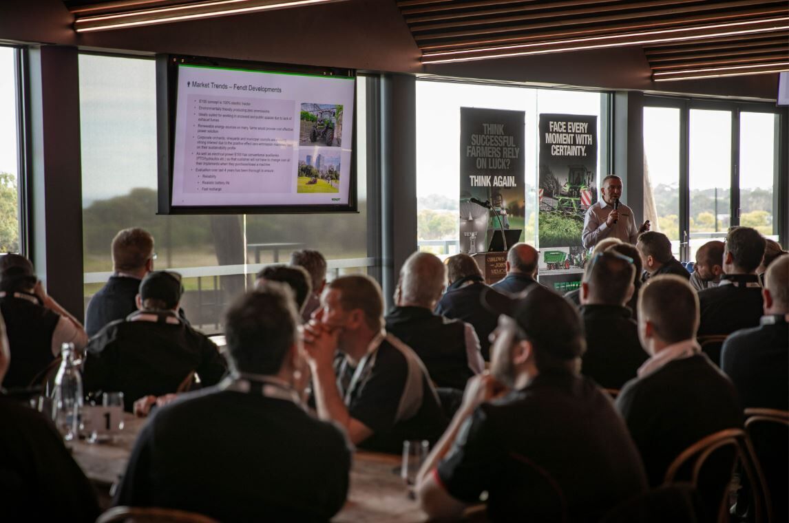 A conference room with attendees seated at tables, a presentation screen, and a speaker at the front.