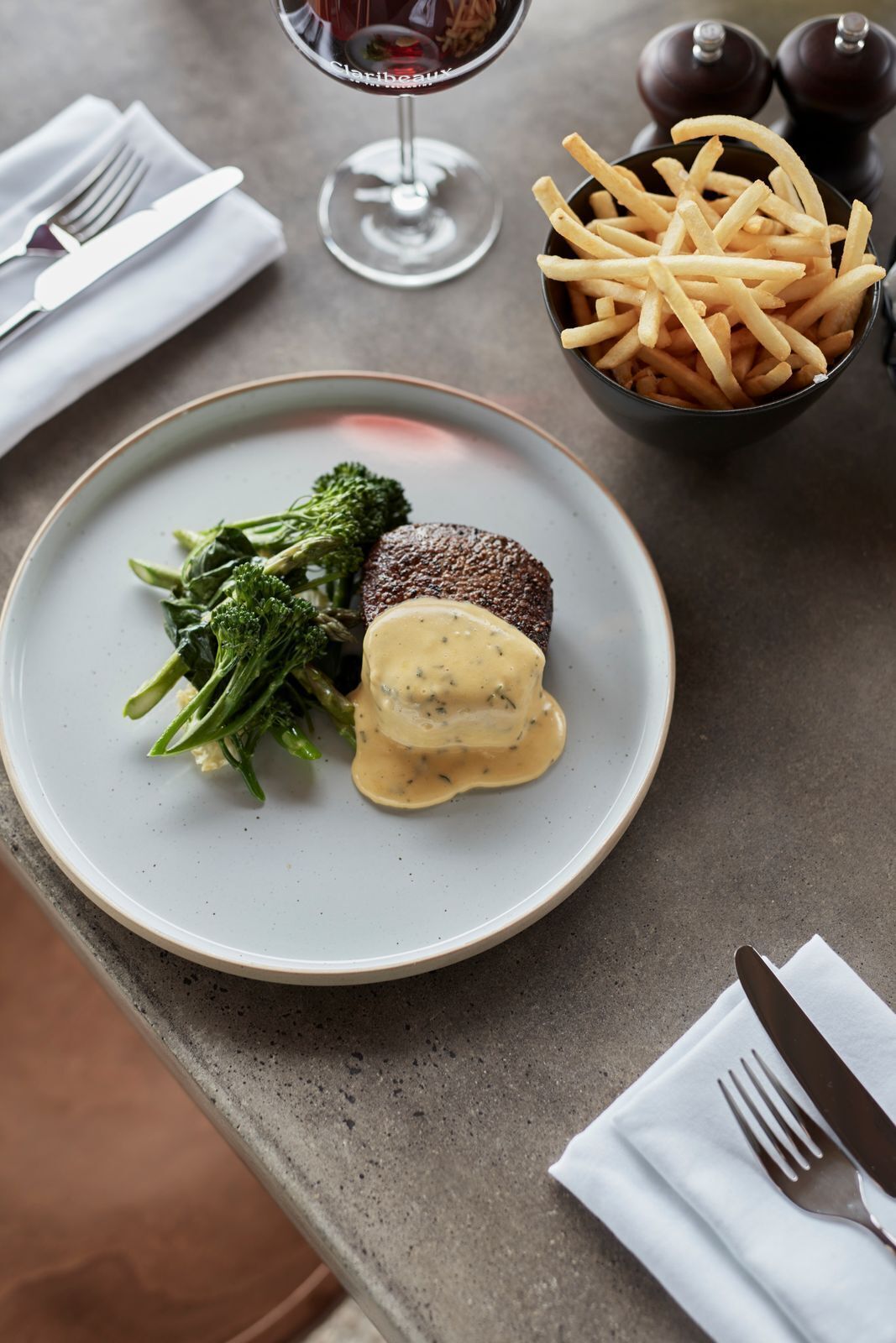 Ein Teller mit einem Steak und Gemüse, ein Glas Rotwein und eine Schüssel Pommes auf einem Tisch.