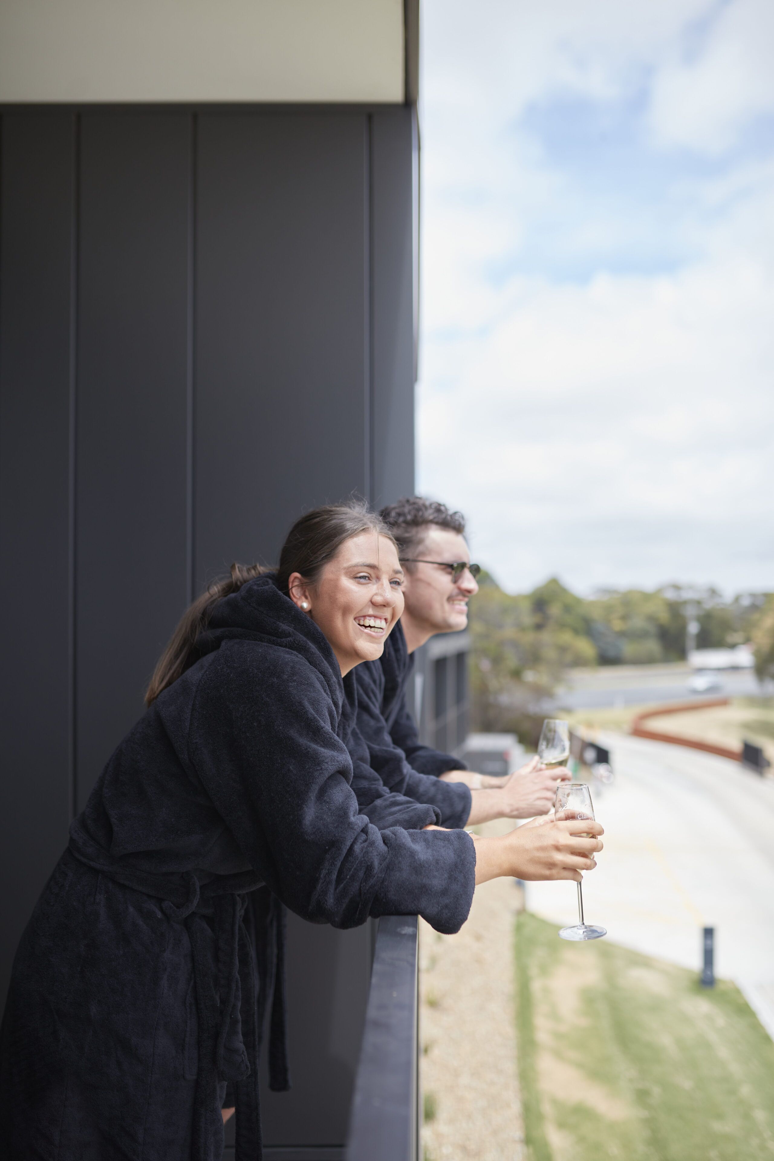 Two people in dark robes leaning on a balcony railing, holding a glass of wine.