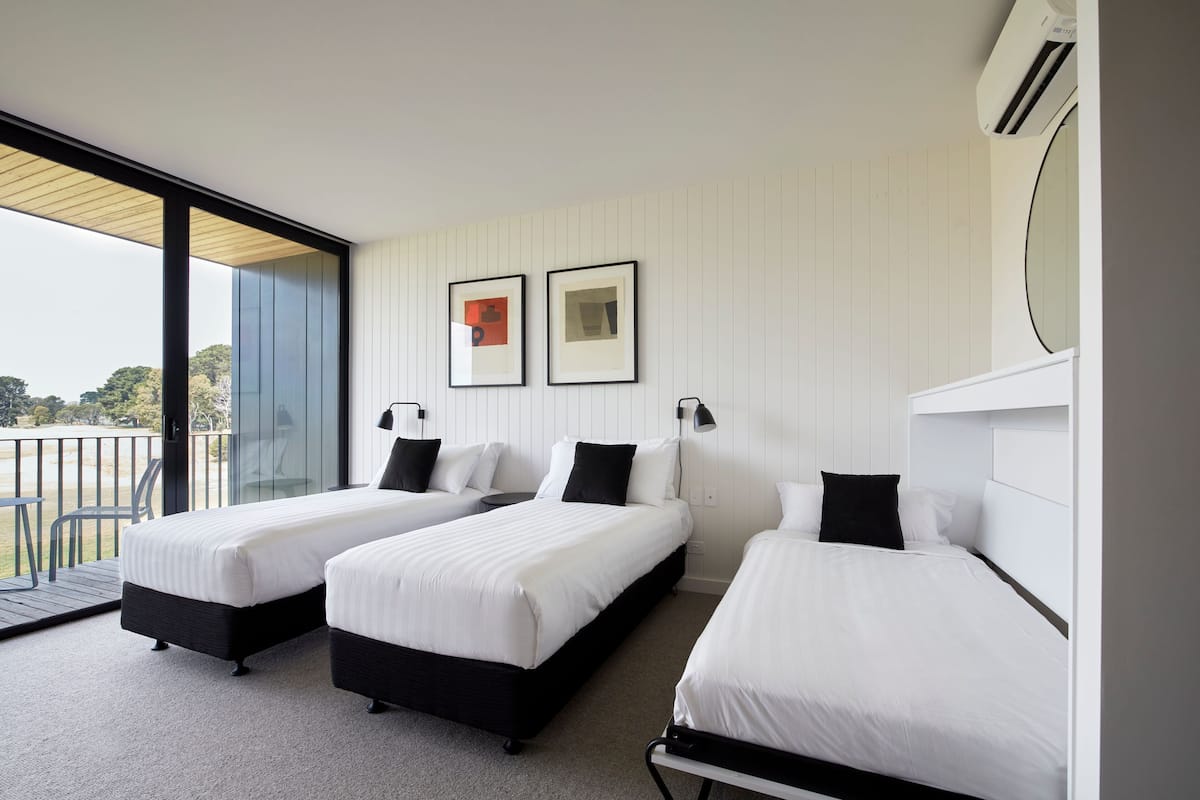 Habitación de hotel moderna con tres camas individuales, un ventanal con vistas al exterior y un balcón.
