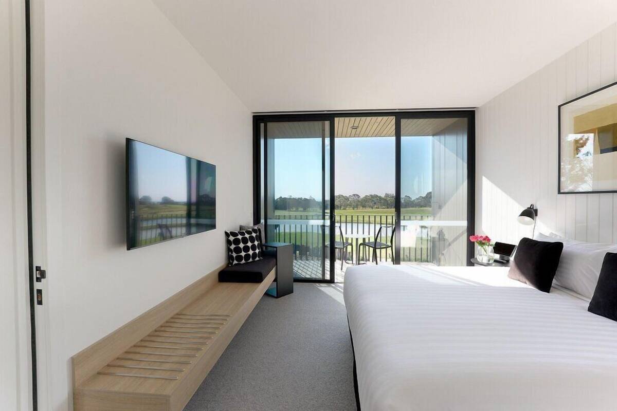 Una habitación de hotel moderna con una cama grande, un televisor de pantalla plana montado en la pared, una zona de estar con un banco y una pequeña mesa, y un balcón con vistas al exterior.