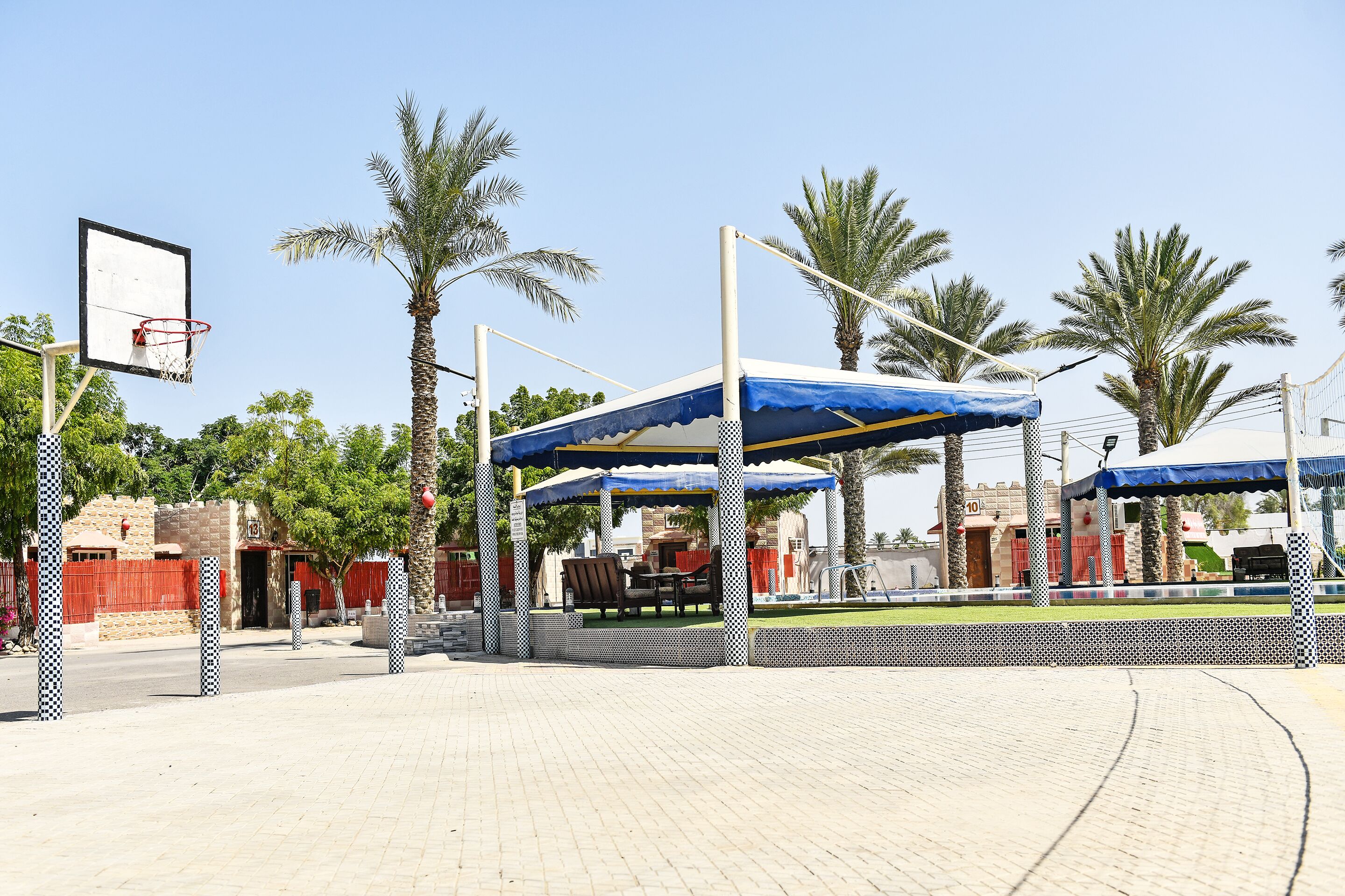 An outdoor basketball court with a blue canopy, palm trees, and a brick building in the background.