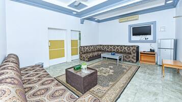 A living room with patterned sofas, a coffee table, a flat-screen TV, and a refrigerator.