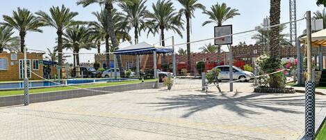 Un espacio exterior empedrado con palmeras, una cancha de baloncesto y un edificio con toldo azul.
