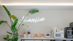 A hotel room with a neon sign reading "Nuber Hotel," a coffee machine, a microwave, and a plant.