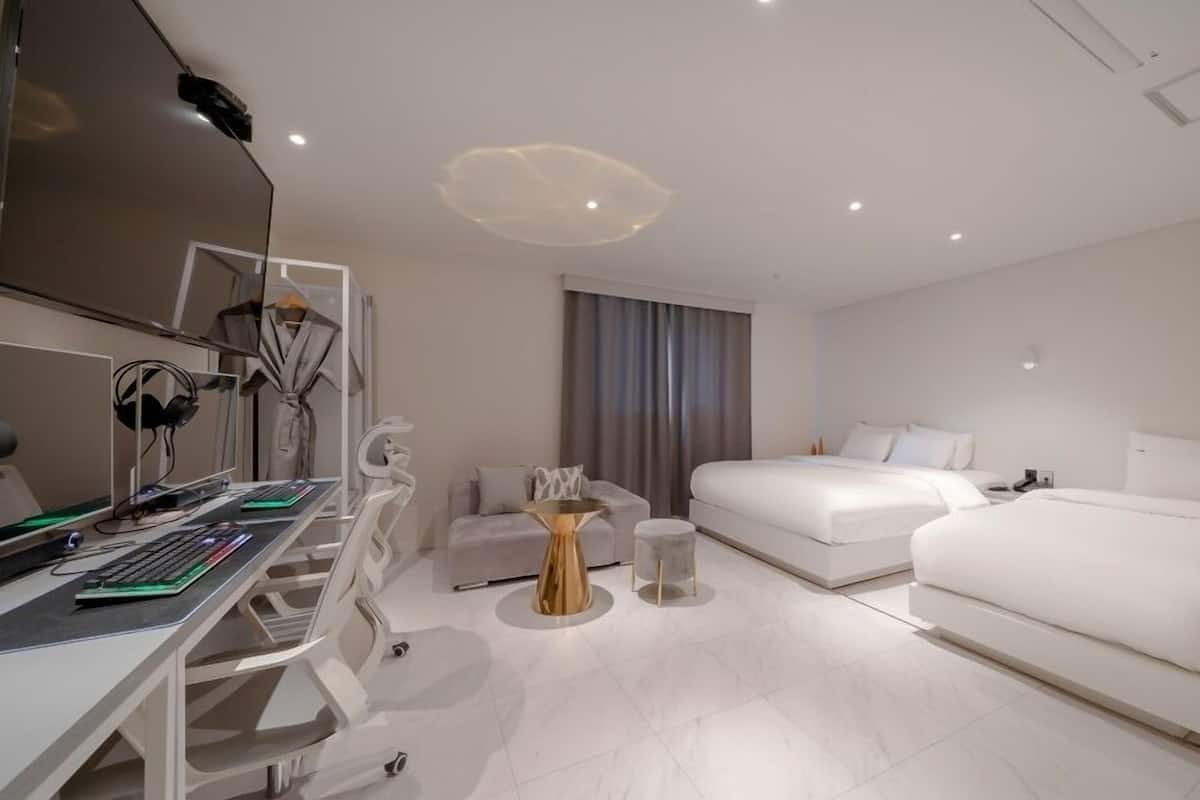 A modern hotel room with a desk, two beds, a vanity with a mirror, and a small seating area.