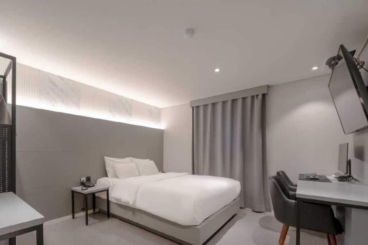 A modern hotel room with a bed, a desk, a chair, and a TV mounted on the wall.