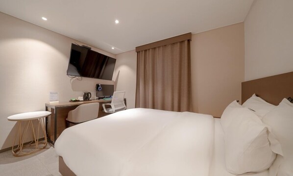 A hotel room with a large bed, a desk with a chair, a flat-screen TV mounted on the wall, and a window with curtains.