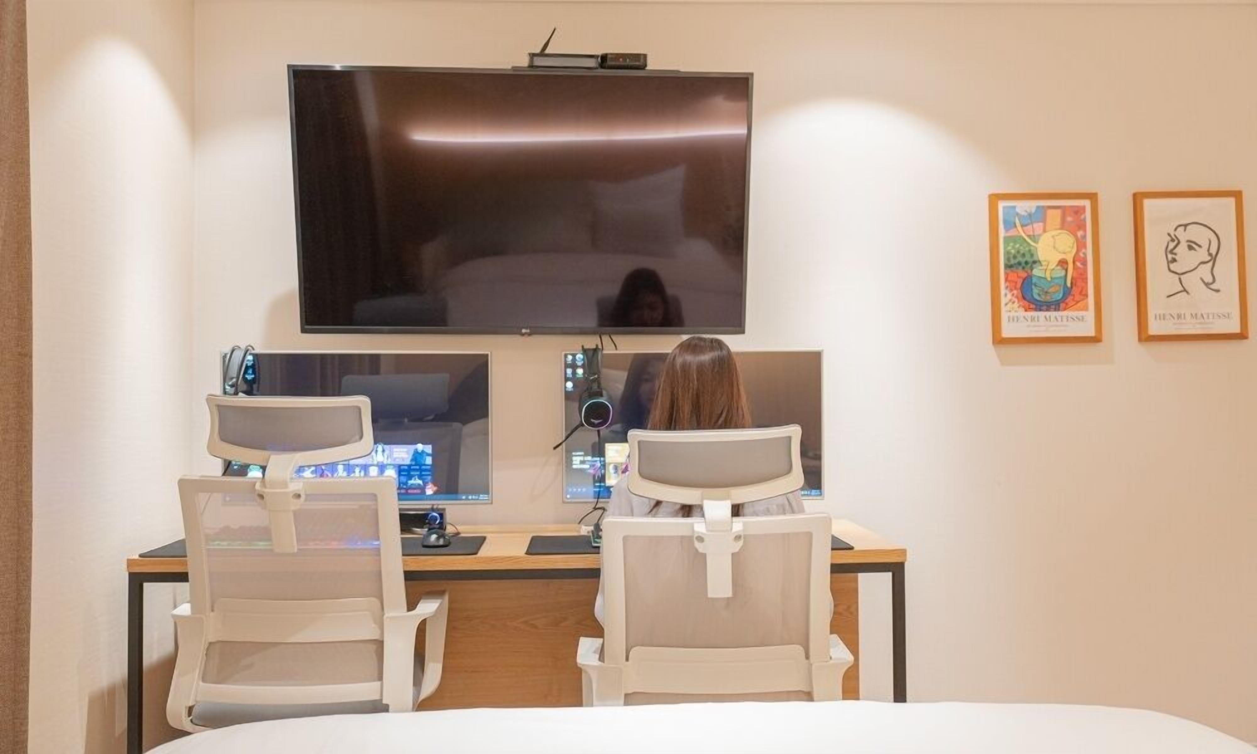 A hotel room with a large flat-screen TV mounted on the wall, two white chairs, a desk with a computer, and two framed artworks on the wall.