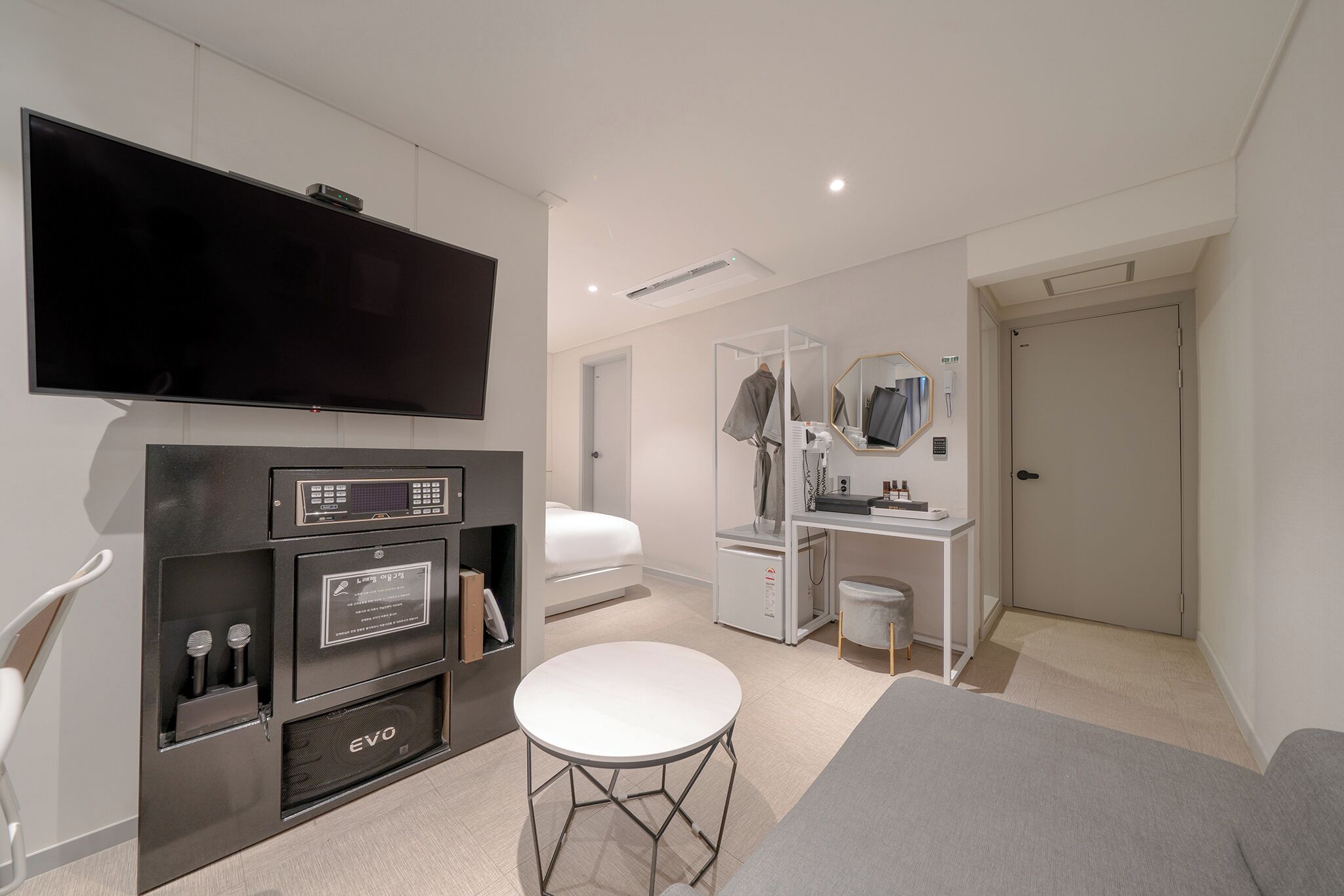 A modern hotel room with a flat-screen TV, a minibar, a desk with a mirror, a vanity, and a bed.