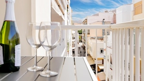 Un balcon avec une bouteille de vin et deux verres, donnant sur un paysage urbain avec des bâtiments et un ciel dégagé.