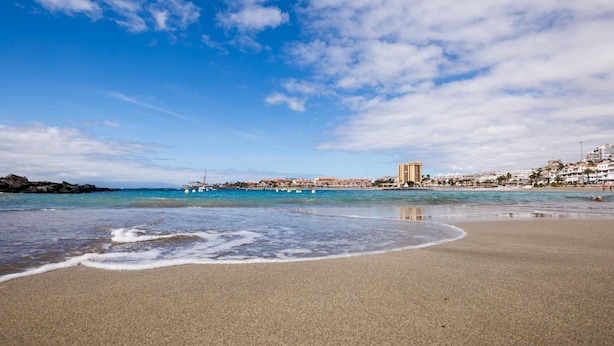 Une plage avec une eau d’un bleu limpide, un rivage sablonneux et des bâtiments en arrière-plan.