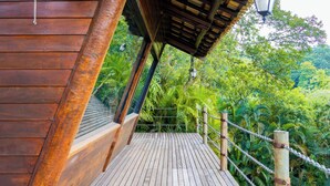 Una terraza de madera con barandilla de cuerda, rodeada de exuberante follaje y una estructura de madera con techo inclinado.