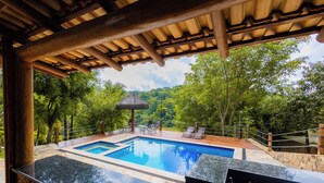 Una piscina con cubierta de madera y sillas de descanso, rodeada de árboles y una estructura con techo de paja.