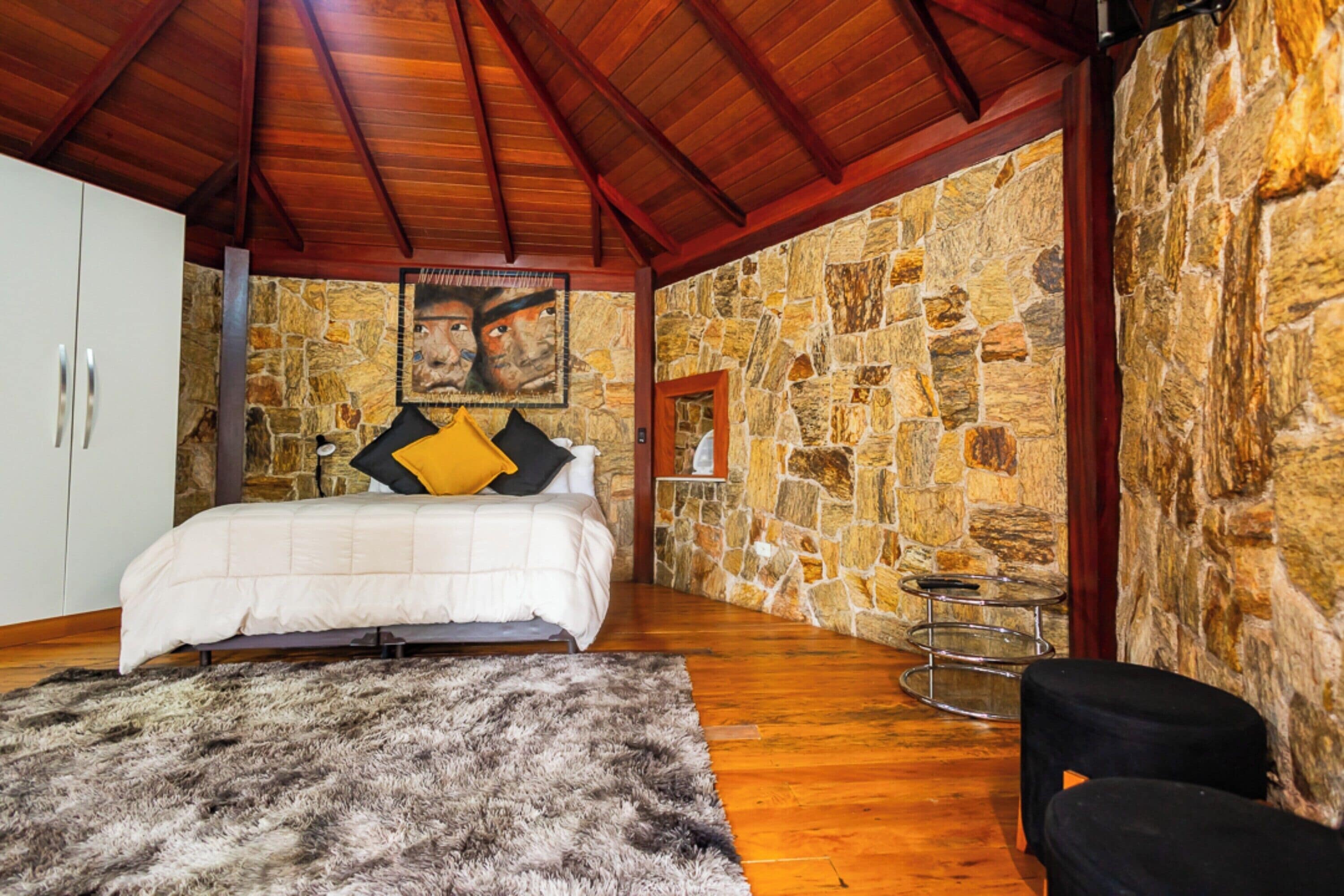 A bedroom with a stone wall, wooden ceiling, a bed, a white wardrobe, a fur rug, and a small round table.