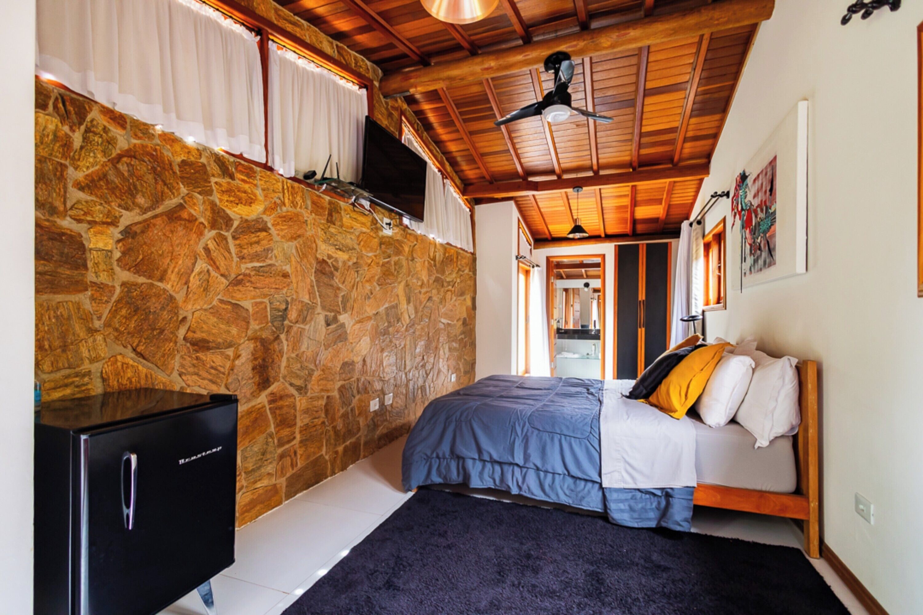 A bedroom with a stone wall, a bed with a blue and white duvet, a black TV on the wall, and a ceiling fan.