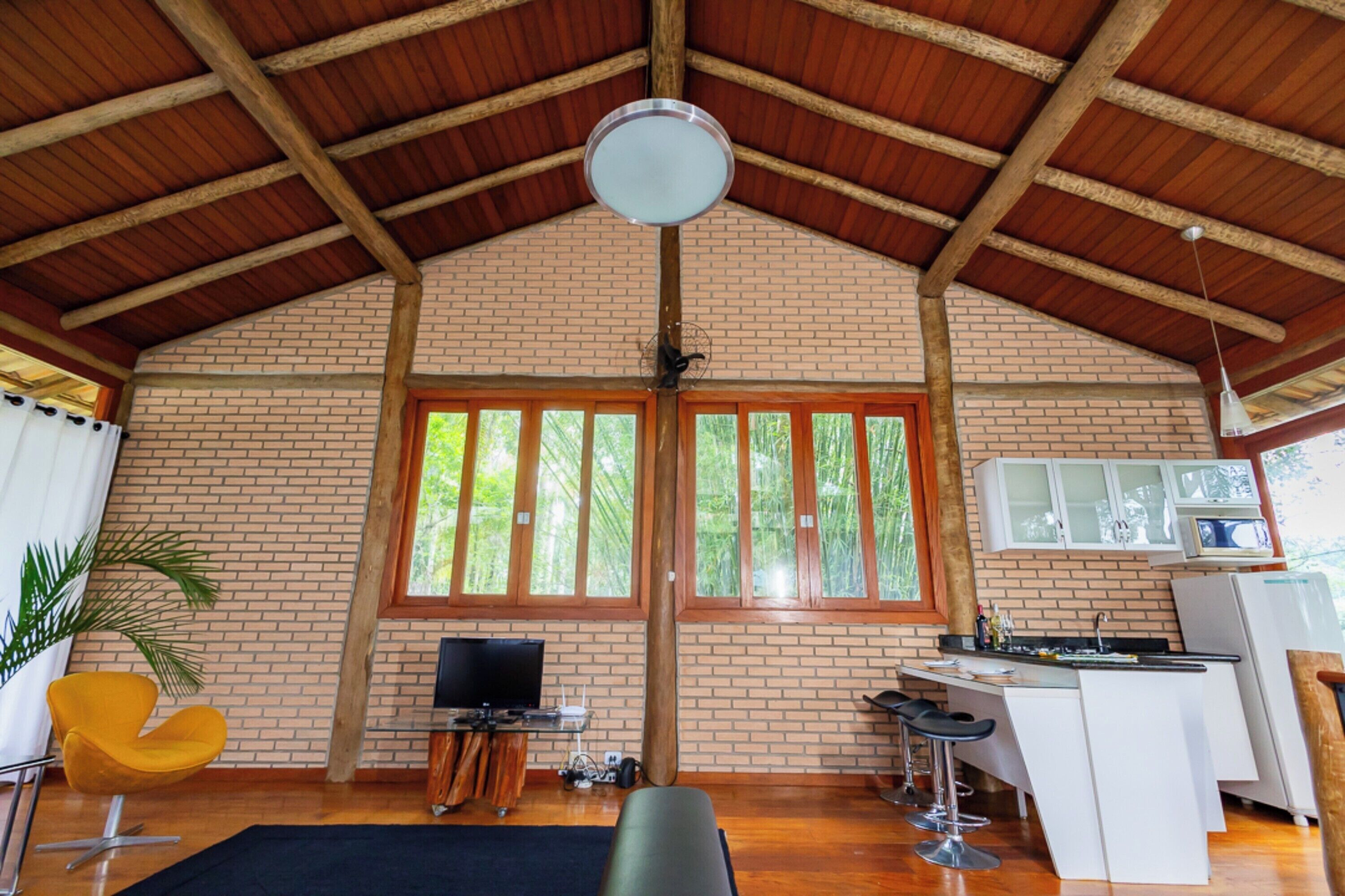 Una habitación amplia con techo alto, paredes de ladrillo, un gran ventanal, televisor, zona de cocina y una silla amarilla.