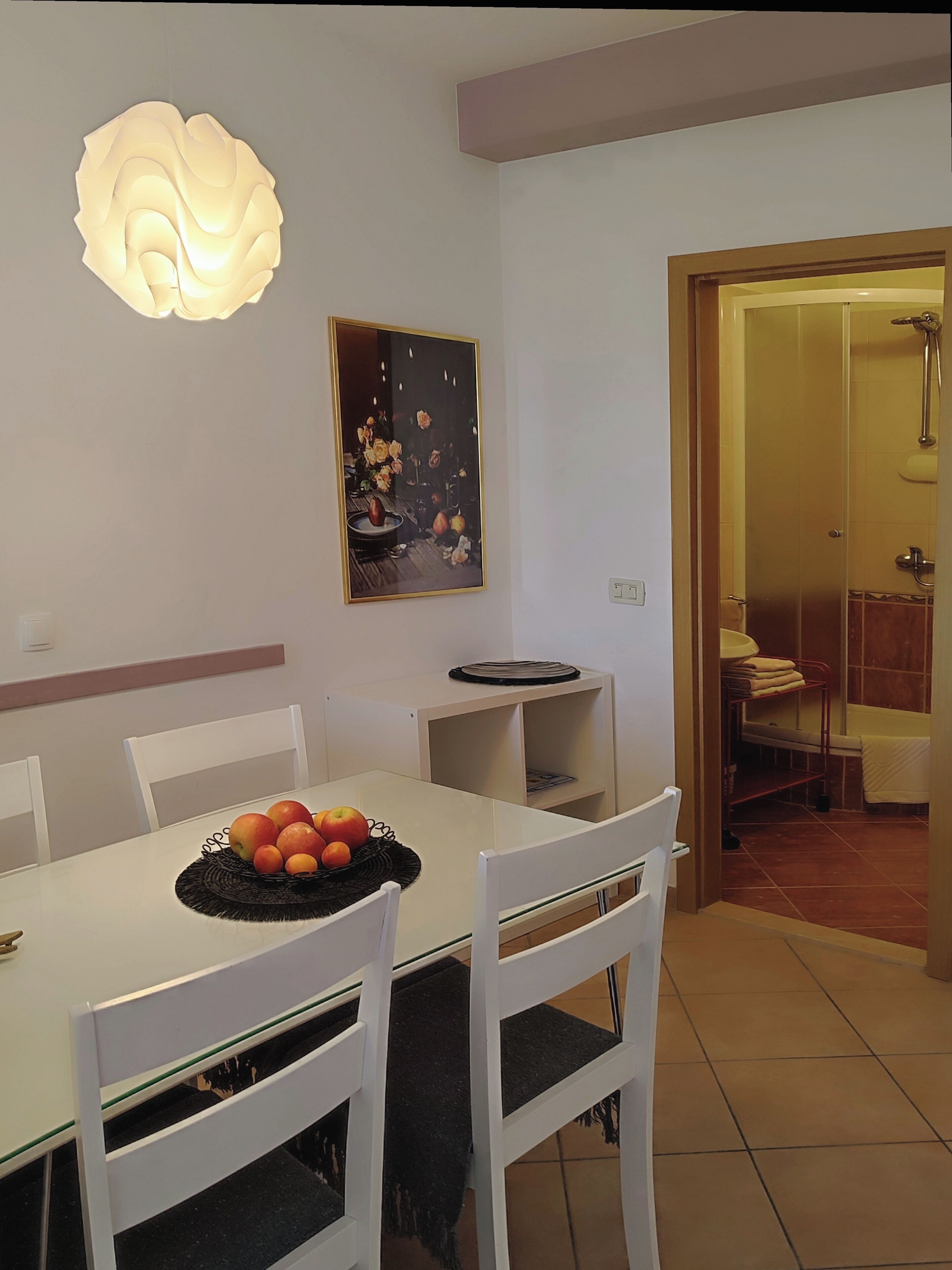 A dining area with a white table, chairs, a fruit basket, a wall-mounted light fixture, and a framed picture.