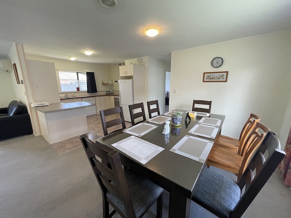 Un comedor con una mesa dispuesta para seis personas, rodeada de sillas de madera. Se ve una cocina al fondo con armarios blancos y un refrigerador.