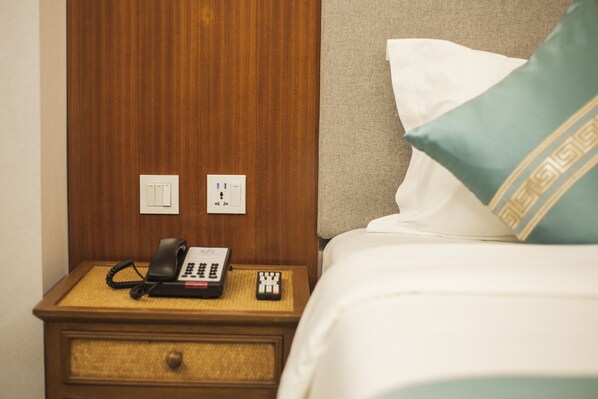 Habitación de hotel con mesita de noche de madera, teléfono, control remoto y cama con almohada blanca y turquesa.