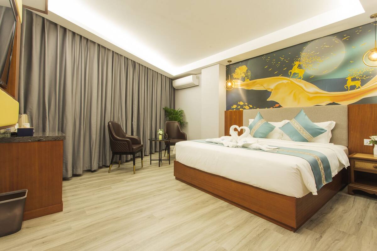 Ein Hotelzimmer mit einem großen Bett, einem Schreibtisch mit Stuhl, einem kleinen Tisch mit Pflanze und einer Wandmalerei.