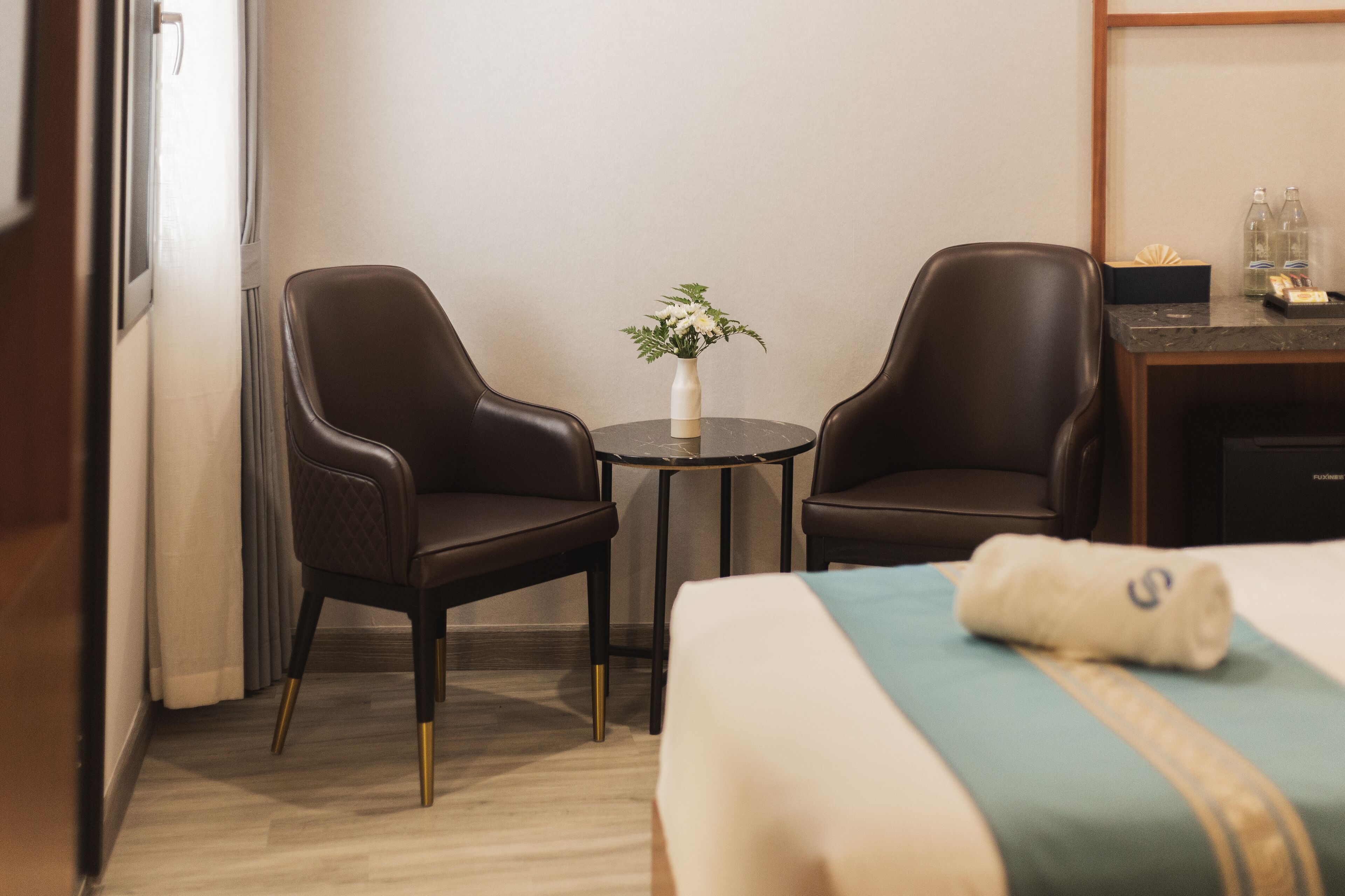 Ett hotellrum med en säng, två läderfåtöljer, ett litet bord med en vas med blommor och ett skrivbord med flaskor och en näsduksask.