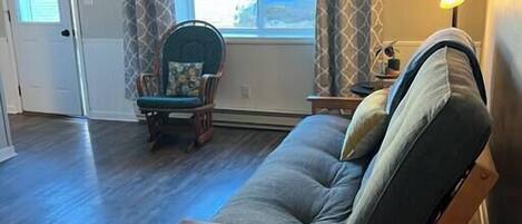 Un salon avec un canapé en bois, un fauteuil, une lampe et une vue sur l’extérieur à travers une fenêtre ornée de rideaux.