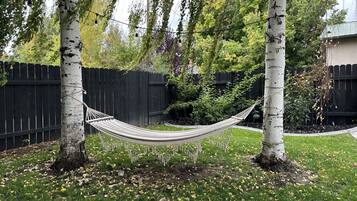 Eine Hängematze zwischen zwei Birken in einem Garten mit Holzlattenzaun und blühenden Pflanzen.