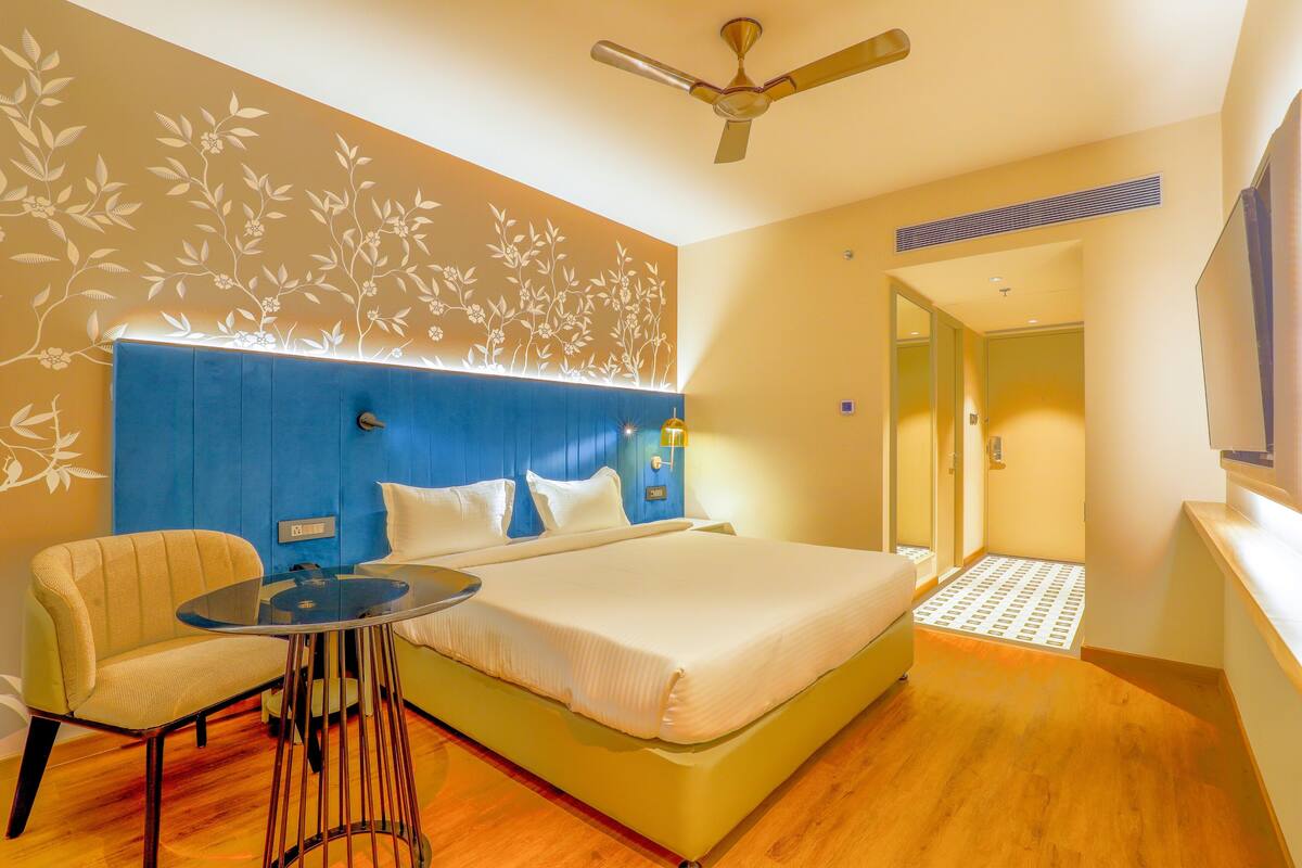 Habitación de hotel con cama, una silla, una mesita, ventilador de techo y televisor.