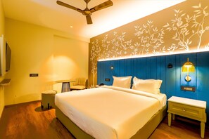 Una habitación de hotel con una cama grande, un ventilador de techo, una lámpara de pared y una pequeña mesa con una silla.