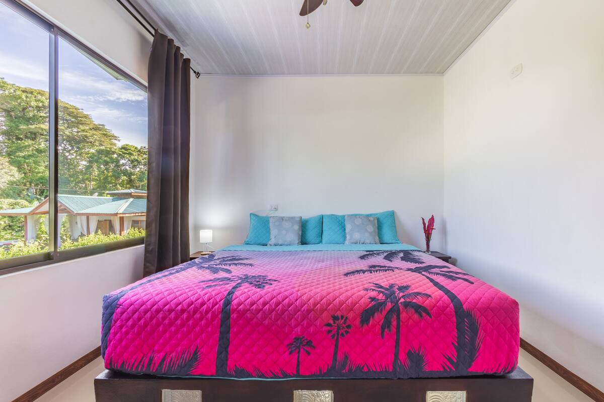 Ein Bett mit einer pink-blauen Steppdecke, ein Fenster mit Blick ins Grüne und ein Deckenventilator.