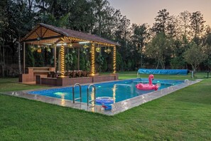 A pool with a lounge area, a covered seating area, and a flamingo float. - The Orchard Farm, Manesar- Luxury Pet- Friendly Farm with private pool & Jacuzzi (Para)