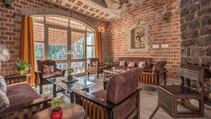 A cozy living room with brick walls, a large window, and a fireplace. - The Orchard Farm, Manesar- Luxury Pet- Friendly Farm with private pool & Jacuzzi (Para)