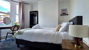 Una cama bien hecha con ropa blanca, una mesita de noche con lámpara, una silla, un escritorio con silla, un perchero y una ventana con cortinas.