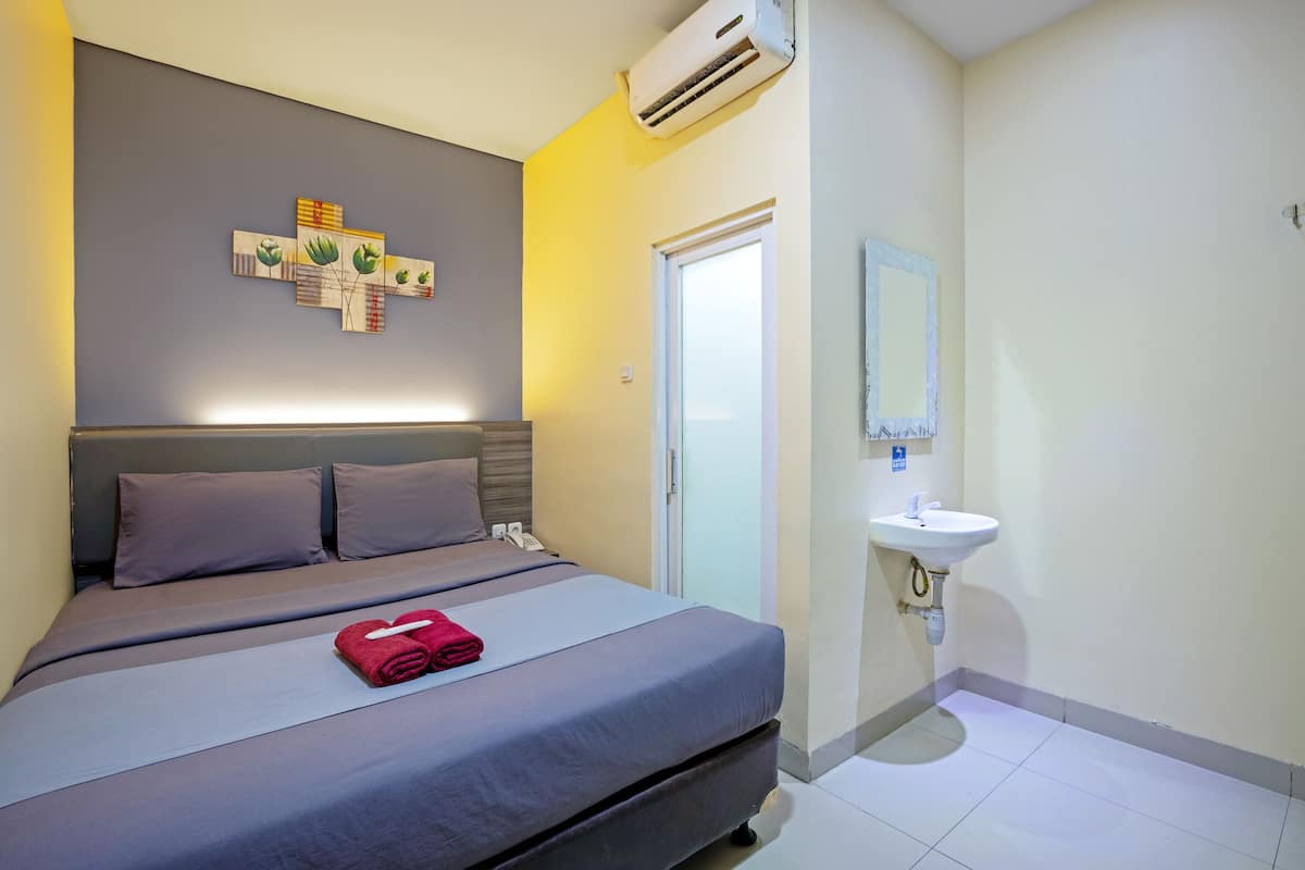 A hotel room with a bed, a wall-mounted air conditioner, a bedside telephone, and a bathroom with a sink and mirror.